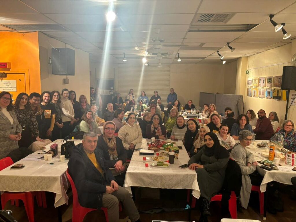 A room full of smiling people looking at the camera while sitting around tables, a number of people are standing to the left of the photo