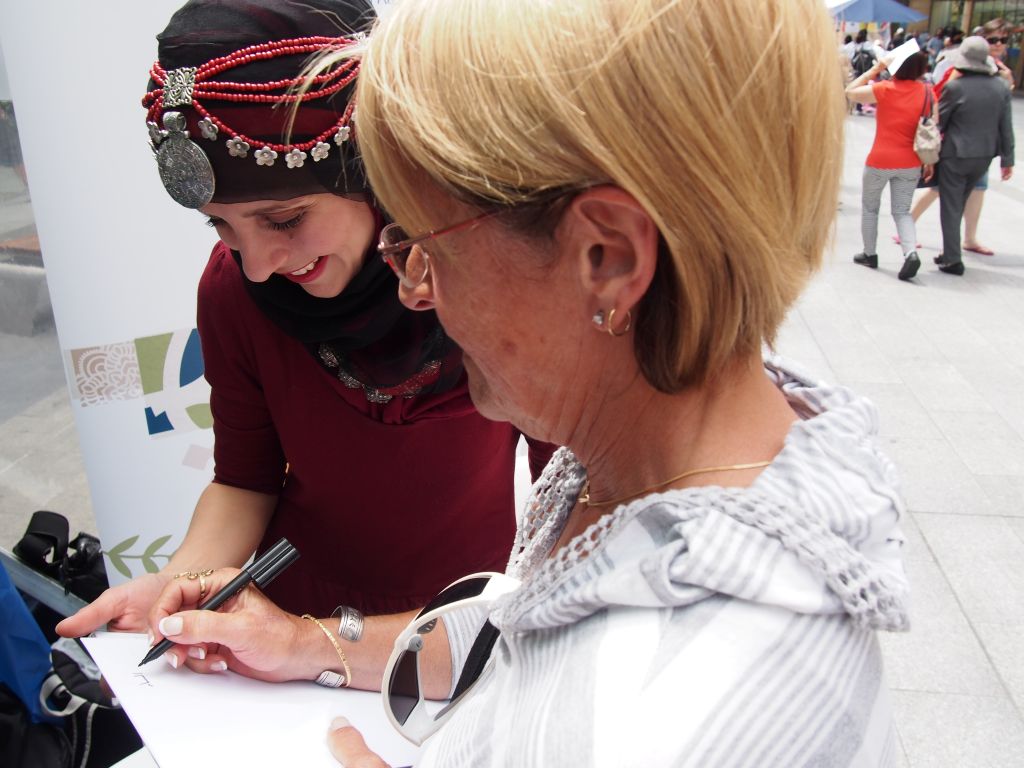 Two women writing on a pad. One woman is wearing a traditional Arabic head dress with attached jewellery